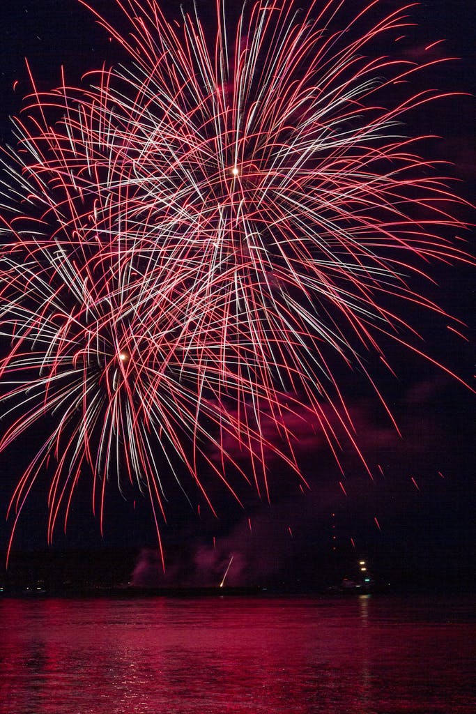 Stunning red fireworks burst over a serene body of water during a night festival celebration.
