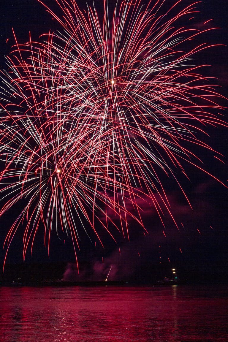 Stunning red fireworks burst over a serene body of water during a night festival celebration.