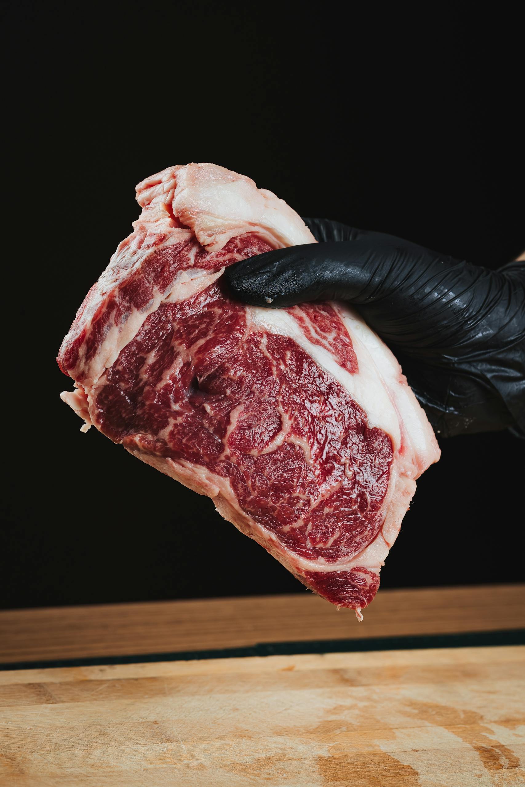Close-up of a raw ribeye steak held by a gloved hand on a wooden surface.